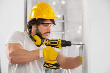 Kaslı bir miğfer takan ve gözlüklü biri, fotokopi alanı olan yüksek tuşlu bir iç mekanda pille çalışan bir matkapla duvarda delik açıyor.