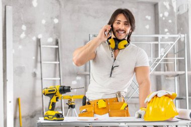 İnşaatçı, mimar ya da inşaat mühendisi, bina planının üzerinde çalışıyor ve kameraya gülümserken cep telefonuyla konuşuyor.