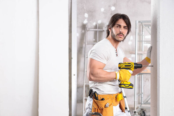 Handsome muscular builder wearing tool belt working with a drill in a new house under construction with copy space