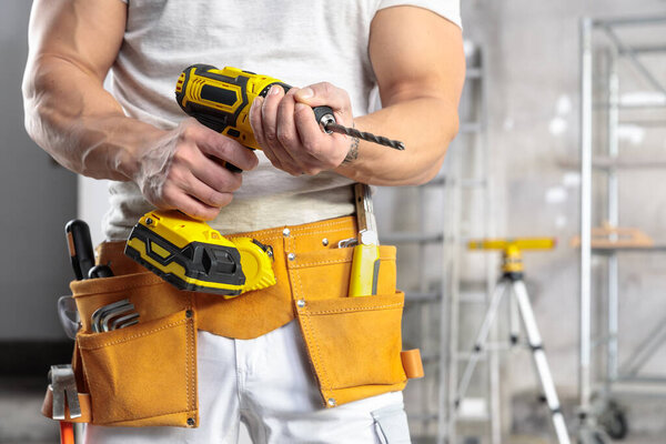 Muscular builder wearing a tool belt holding a battery operated drill in a room under construction in a close up on his hands