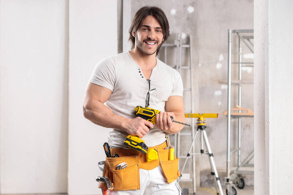 Muscular builder with a friendly smile standing in a high key room under construction holding a battery operated drill with copy space