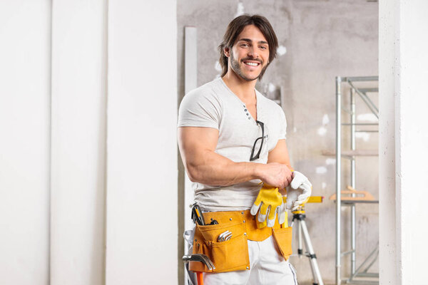 Friendly builder wearing a tool belt and gloves standing smiling at the camera in a high key room under construction with copy space