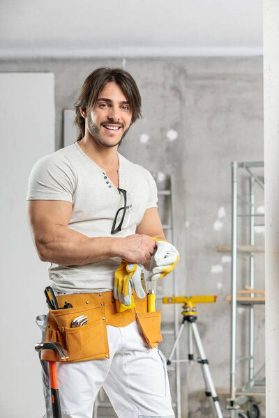 Friendly builder wearing a tool belt and gloves standing smiling at the camera in a high key room under construction with copy space