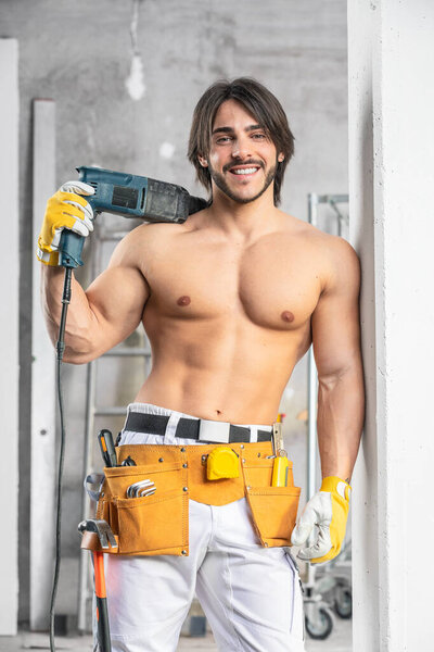 Smiling sexy muscular builder posing shirtless with a toll belt around his hips and power drill slung over his shoulder inside a room under construction