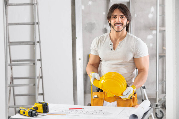 Smiling builder or structural engineer on site standing holding his hardhat over a blueprint of the building on a workbench with lateral copy space
