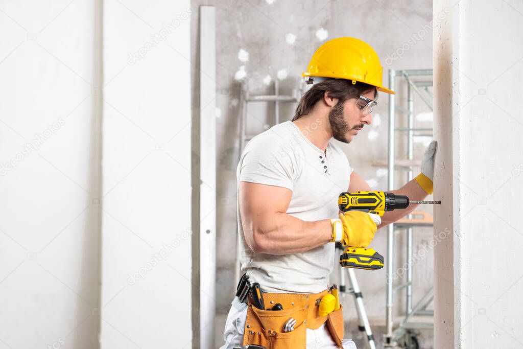 Handsome muscular builder wearing a hardhat and tool belt working with a drill in a new house under construction with copy space