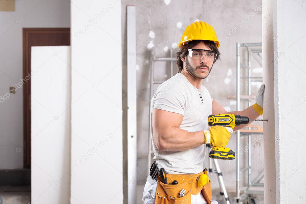 Handsome muscular builder wearing a hardhat and tool belt working with a drill in a new house under construction with copy space