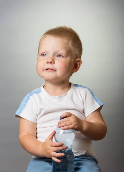 Nachdenklicher kleiner Junge mit einer Plastikflasche Trinkwasser auf grauem Hintergrund — Stockfoto