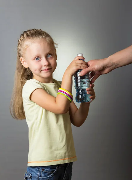 Blonde girl takes plastic bottle with water from her mother on gray background — Stock Photo, Image