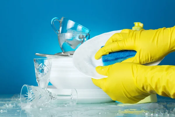 Sarı eldivenli eller açık mavi arka planda mavi sünger tabağını yıkar. — Stok fotoğraf
