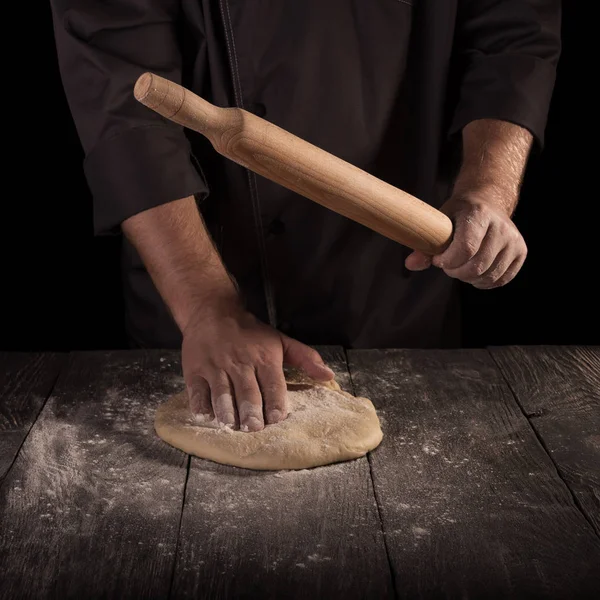 Chef man in dark jacket roll out the dough isolated on black background — Stock Photo, Image