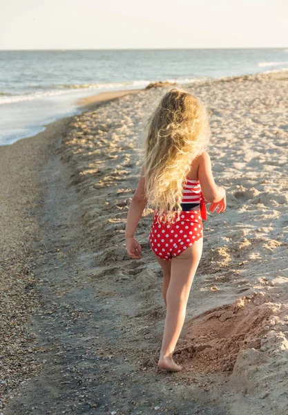 Lille mørkhåret pige i en rød badedragt går langs varm, solrig strand - Stock-foto