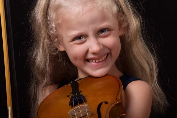 Учусь играть на скрипке. Улыбающаяся маленькая девочка держит скрипку в руках на темном фоне — стоковое фото