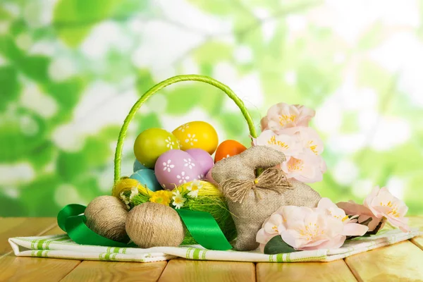 Färgade påskägg i korg, sammanflätade snoddar, kanin, blommor — Stockfoto