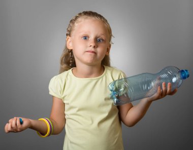 Şaşırmış küçük kız elinde boş bir şişeyle, gri üzerinde duruyor.