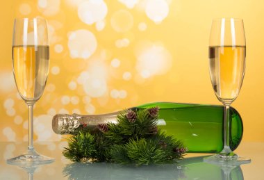 New year champagne bottle lies on the table near pine branches with pine cones and two full wine glass, on yellow background