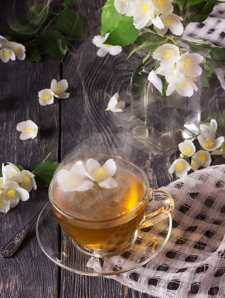 Ατμός πάνω από Κύπελλο ζεστό τσάι, γιασεμί λουλούδια είναι διάσπαρτα σε γκρι επιφάνεια — Φωτογραφία Αρχείου