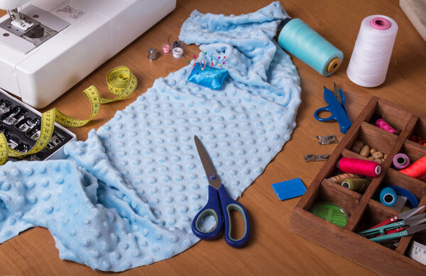 Sewing machine, drawer with accessories, piece of light blue cloth and scissors on desktop