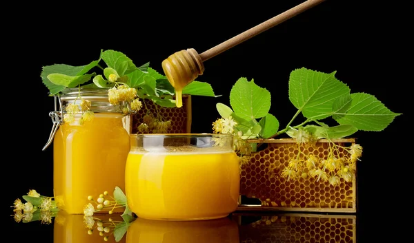 Dois frascos de vidro cheios de mel dourado, favos de mel de abelha e flores de tília isoladas em preto — Fotografia de Stock