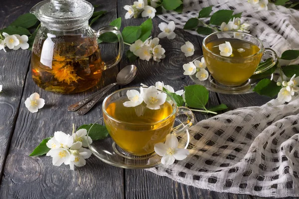 Konvice s bylinkovým čajem, šálky s pitím, roztroušené jasmínové květy na šedém povrchu — Stock fotografie