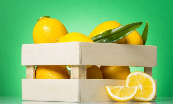 Lemons with leaves in wooden box, next to lemon wedges, on green — Zdjęcie stockowe