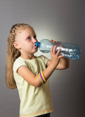 Uzun saçlı küçük kız, gri arka planda şişeden su içiyor.