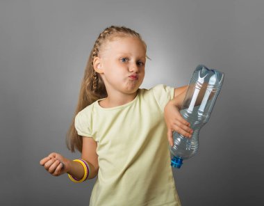 Hayal kırıklığına uğramış küçük bir kız, gri arka planda, içme suyunun altından boş bir plastik şişe tutuyor.