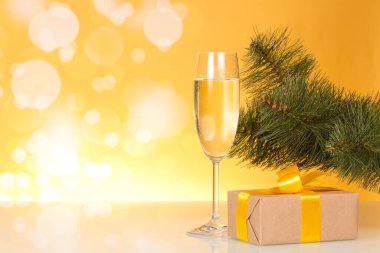 Noel ağacı dalı, bir kadeh şampanya ve parlak sarı arka planda güzel bir fiyonklu hediye kutusu.