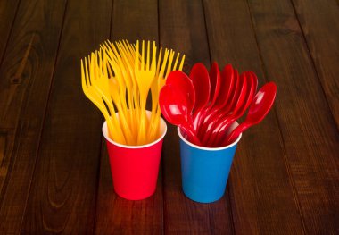 Tek kullanımlık parlak kaşıklar ve çatallar plastik bardaklarda, kahverengi arka planda ahşap masa