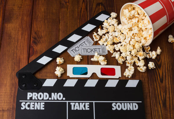 Movie tickets, 3D glasses, paper box spilled popcorn and movie clapper on a wooden background