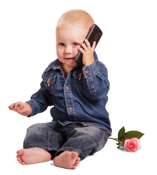 一个穿着蓝色衬衫和牛仔裤的小男孩坐在靠近耳朵的地方 手里拿着一部手机 被白色的背景隔离了起来 — 图库照片