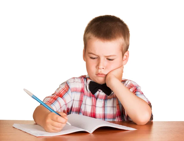 白い背景に隔離されたノートブックに机の上に座っている彼の手にペンを持つ悲しい学童 — ストック写真