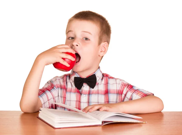Słodki Chłopiec Jedząc Jabłko Siedząc Przy Stole Nad Książką Odizolowany — Zdjęcie stockowe