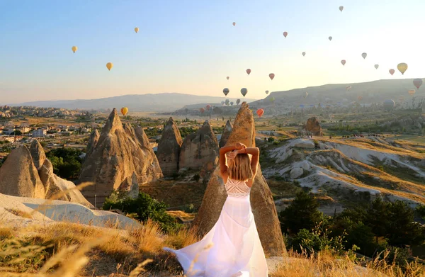 長い白いドレスを着たセクシーな若い女の子の旅行者は カッパドキアトルコの風船を背景に山の上に立っています トルコを参照 素晴らしい眺め — ストック写真