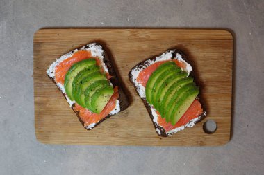 Somon ve avokado tostu krem peynirli ve çavdar ekmeği ahşap masa üzerinde gri arka planda..