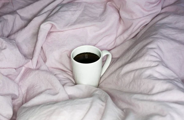 Morning coffee mug in pink bed sheet background. Cotton textile waved.