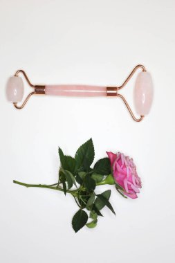 Beyaz arka planda yüz silindiri ve pembe gül. Güzellik aleti yaşlanmayı önlüyor. Yüz tedavi aygıtı.