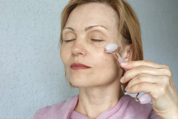 Mature woman using pink face roller. Skin treatment.