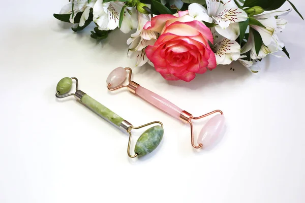 Jade face roller for beauty facial massage therapy. Beauty tools pink and green face roller  on flowers background. Face treatment. Home spa.
