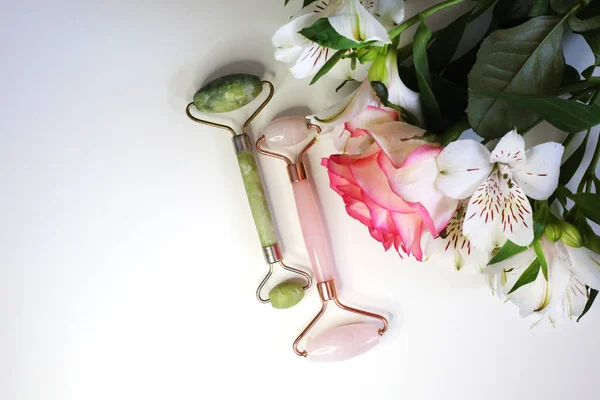 玉面辊用于美容面部按摩治疗 漂亮的工具 粉色和绿色的脸辊在花朵的背景上 面部治疗 家庭温泉 — 图库照片