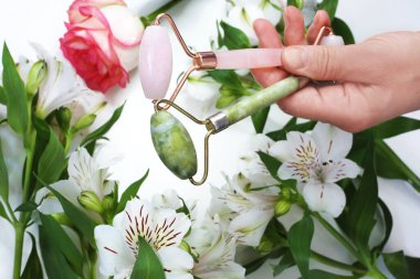 El ele tutuşan pembe ve yeşil yüz silindirleri. Çiçekli bahar arka planında güzellik aletleri yüz silindiri. Yüz bakımı. Ev kaplıcası. 