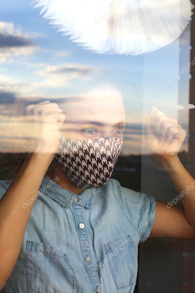 Sad woman standing near window and wearing medical face mask during quarantine and self isolation. Home quarantine concept. Prevention COVID-19 corona virus. Woman looking through the window.