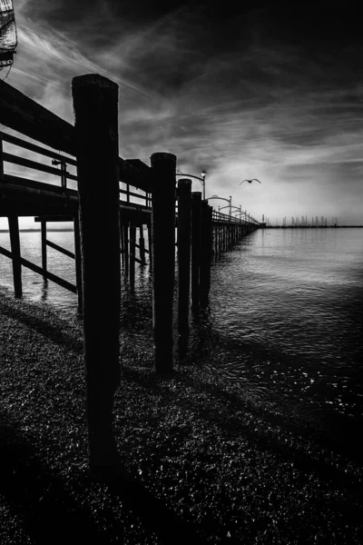 Vertical greyscale shot of a wooden dock with columns on the lake under the beautiful storm clouds — Stock Photo, Image