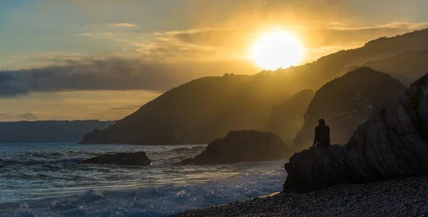 Person sitting on the rock on a beach in front of the sea with the sun rising over a cliff — ストック写真