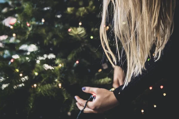 Блондинка в черном свитере украшает елку огнями — стоковое фото