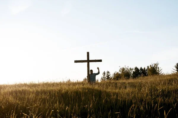 Homem de pé perto de uma cruz de madeira com a mão para cima em direção ao céu em um campo gramado — Fotografia de Stock