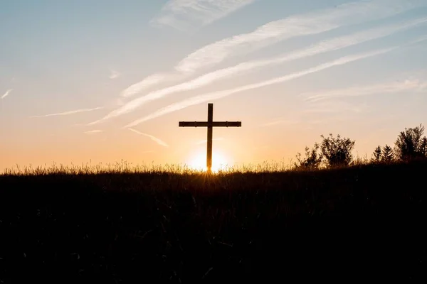 Silhueta de uma cruz de madeira em uma colina gramada com o sol brilhando no fundo — Fotografia de Stock