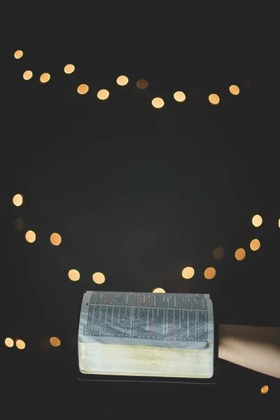 Vertikální detailní záběr osoby držící otevřenou bibli s rozmazanými světly v pozadí — Stock fotografie