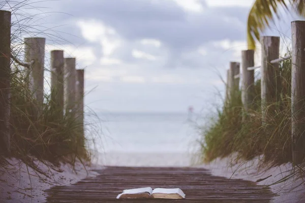 Close-up tiro de uma bíblia aberta em um caminho de madeira em direção à praia com um fundo borrado — Fotografia de Stock