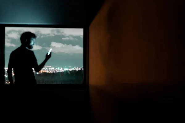 Silueta de un hombre mirando su teléfono con la ciudad de la noche en el fondo — Foto de Stock